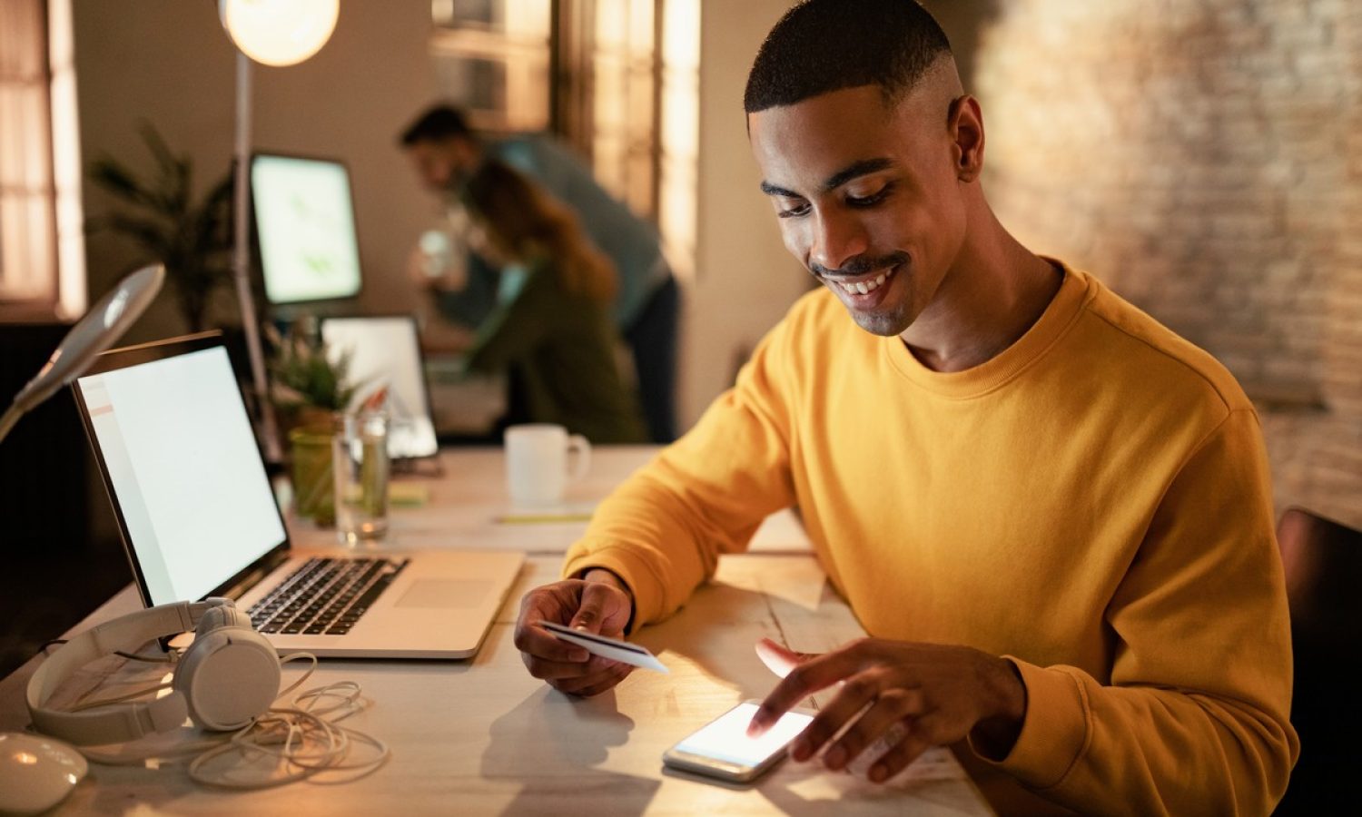 Happy African American entrepreneur using mobile phone and credit card from online payments while working at night in the office.