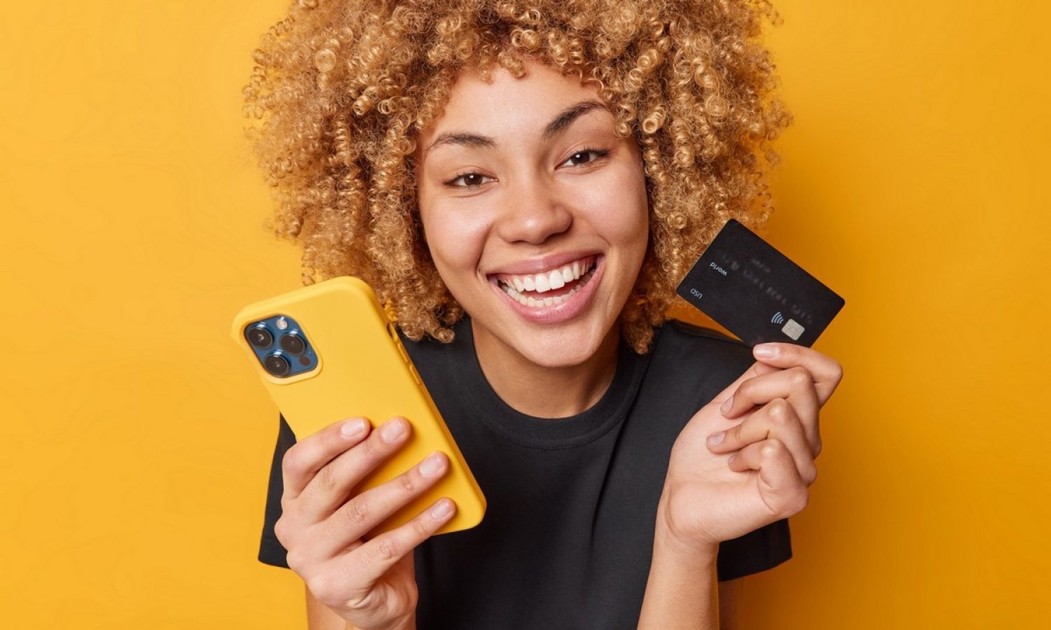 Happy curly female shopper enjoys easy paying for goods online smiles broadly shows white teeth uses mobile phone and credit card dressed in casual black t shirt isolated over yellow background